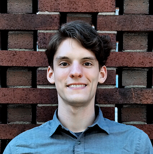 Close up of Alex standing in front of a red brick wall wearing a medium gray button down shirt.